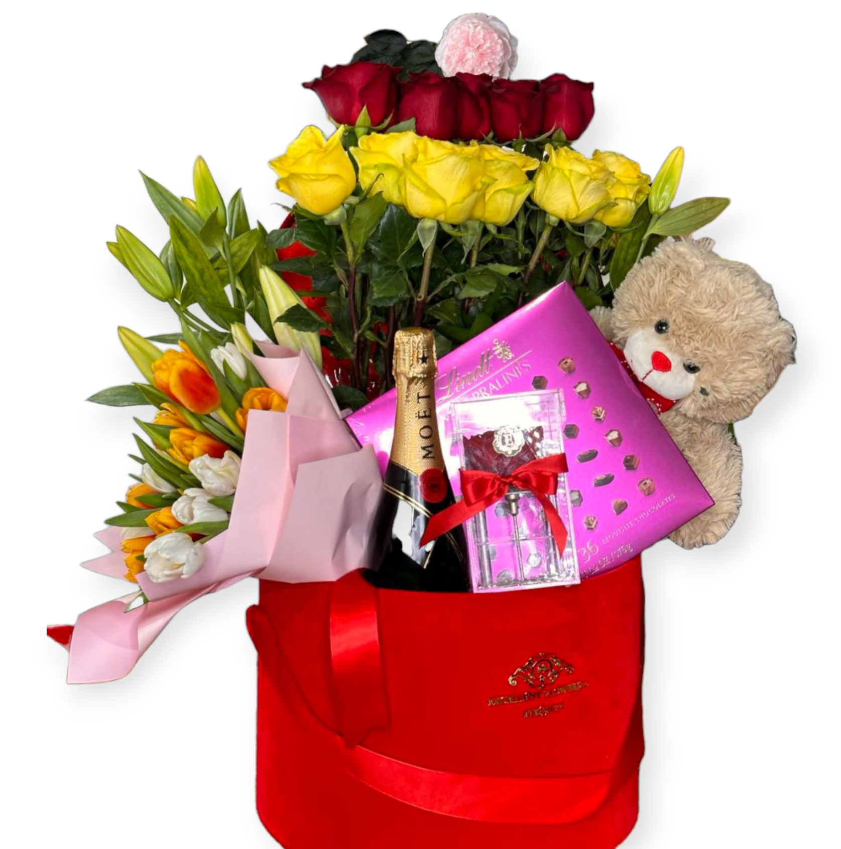 A floral bouquet featuring a mix of red and yellow roses, tulips, and a teddy bear, with champagne and chocolates presented in a red gift bag.