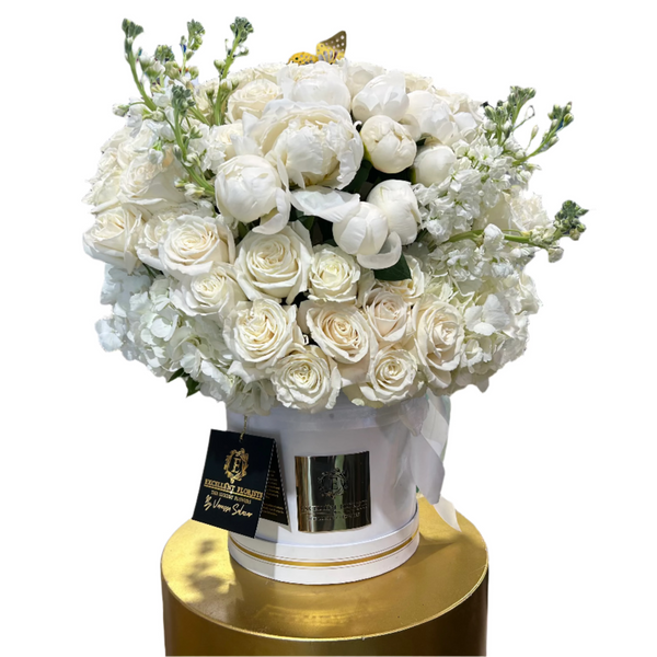 A luxurious floral arrangement featuring premium white roses, peonies, hydrangeas, and stock flowers in a white round box with gold accents, presented on a gold pedestal.