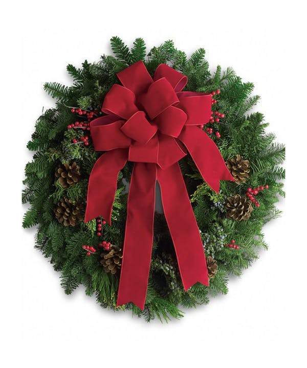 A round holiday wreath decoration featuring green noble fir and juniper branches, red velvet bow, pinecones, and berry sprays.