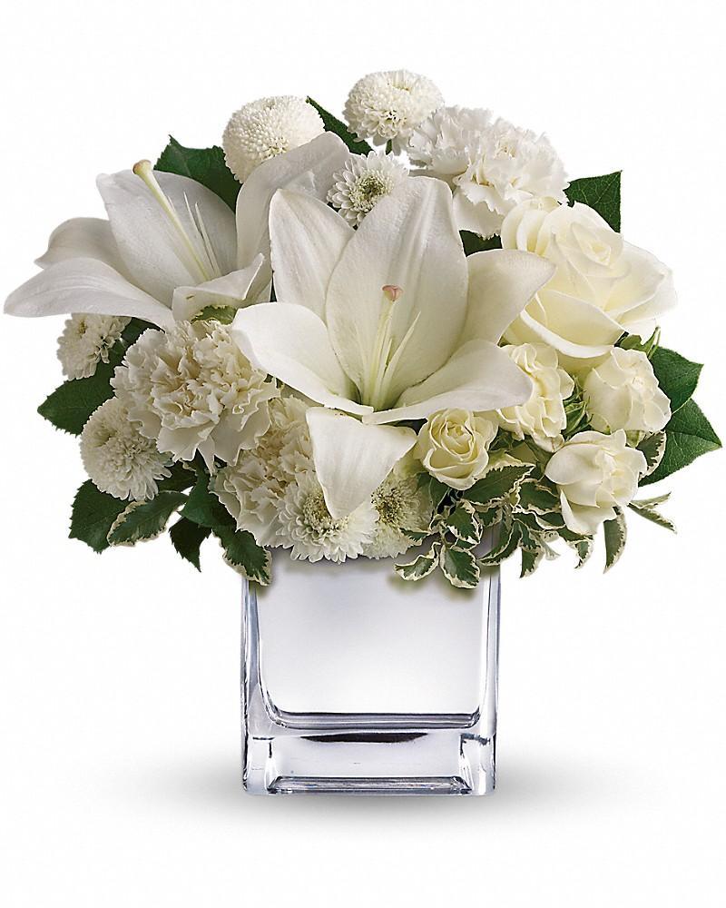 A bouquet of white flowers including roses and lilies, arranged in a clear cube vase.